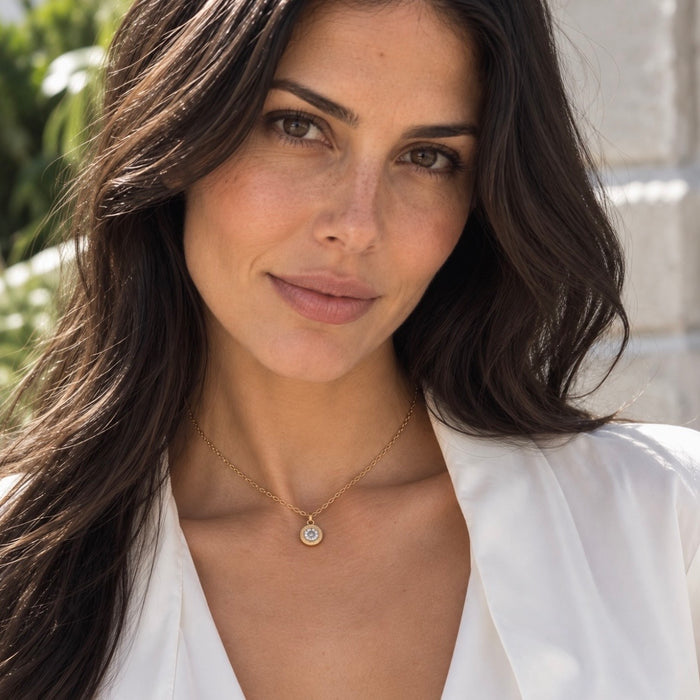 A close-up portrait of a woman with long dark brown hair wearing a delicate gold chain necklace with a small round pendant, paired with a white blouse. The background is a softly blurred outdoor setting.