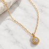 A gold chain necklace with a round bezel-set clear gemstone pendant, displayed on a white marble surface next to a white rock.