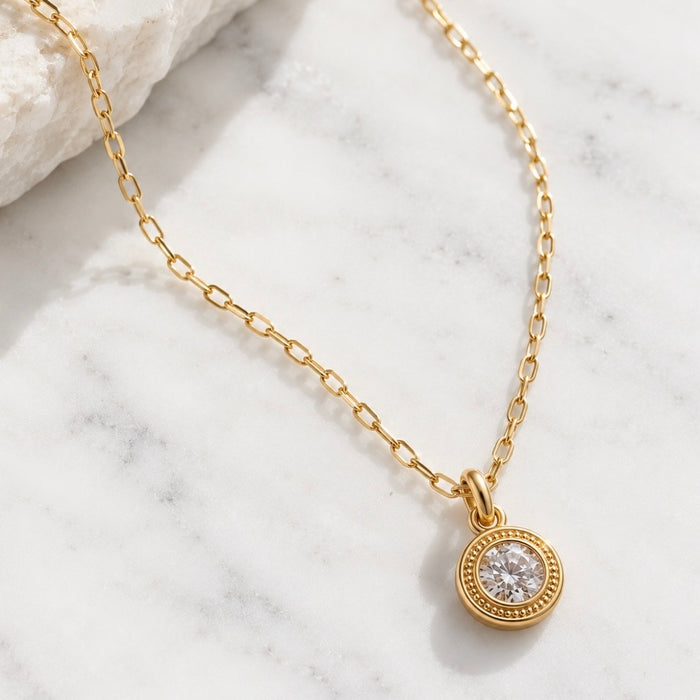 A gold chain necklace with a round bezel-set clear gemstone pendant, displayed on a white marble surface next to a white rock.