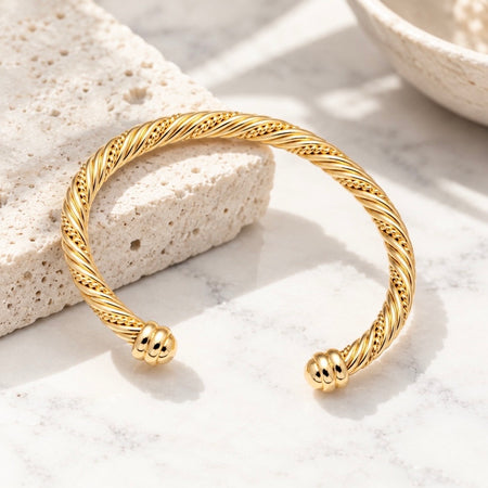 A gold twisted cable open cuff bracelet with beaded ends, displayed on a light marble surface next to a textured stone block.