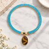 A gold-tone chain tennis bracelet with bright turquoise rectangular links and an oval tiger's eye pendant, displayed on a white marble surface.