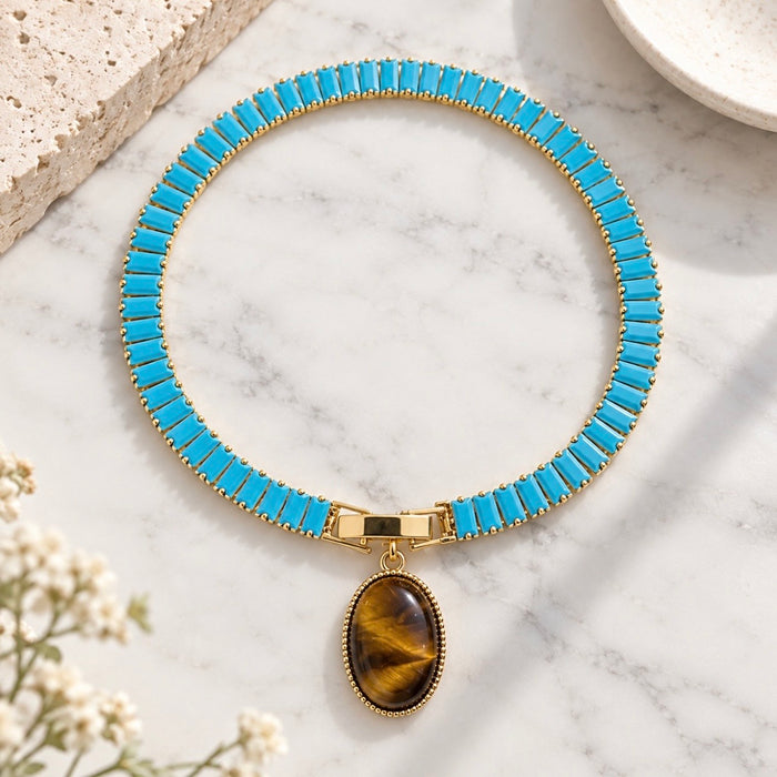 A gold-tone chain tennis bracelet with bright turquoise rectangular links and an oval tiger's eye pendant, displayed on a white marble surface.