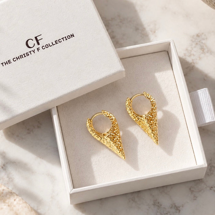 A pair of textured gold hoop earrings displayed in an open white jewelry box, with the branded lid resting beside it on a light marble surface.