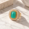 A gold signet-style ring with a large green cabochon center stone and a halo of small clear stones, photographed on a white marble surface with soft sunlight and shadows.