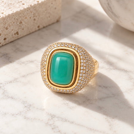 A gold signet-style ring with a large green cabochon center stone and a halo of small clear stones, photographed on a white marble surface with soft sunlight and shadows.