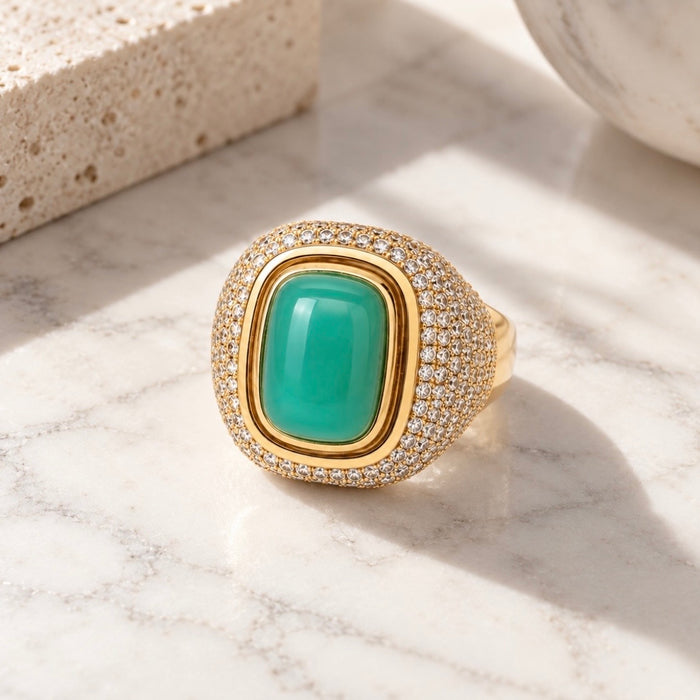 A gold signet-style ring with a large green cabochon center stone and a halo of small clear stones, photographed on a white marble surface with soft sunlight and shadows.