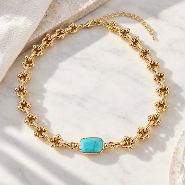A gold chain necklace with a central rectangular turquoise stone, displayed on a white marble surface with soft shadows.