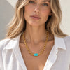 A close-up of a woman wearing a gold chain necklace with a rectangular turquoise stone pendant, styled with a white linen shirt against a neutral light background.