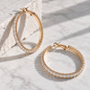 Gold hoop earrings paved with clear rectangular gemstones, displayed on a white marble surface next to a textured white stone block.