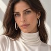 A close-up portrait of a woman wearing large silver-toned hoop earrings studded with small clear crystals, paired with a cream turtleneck top against a softly lit light background.