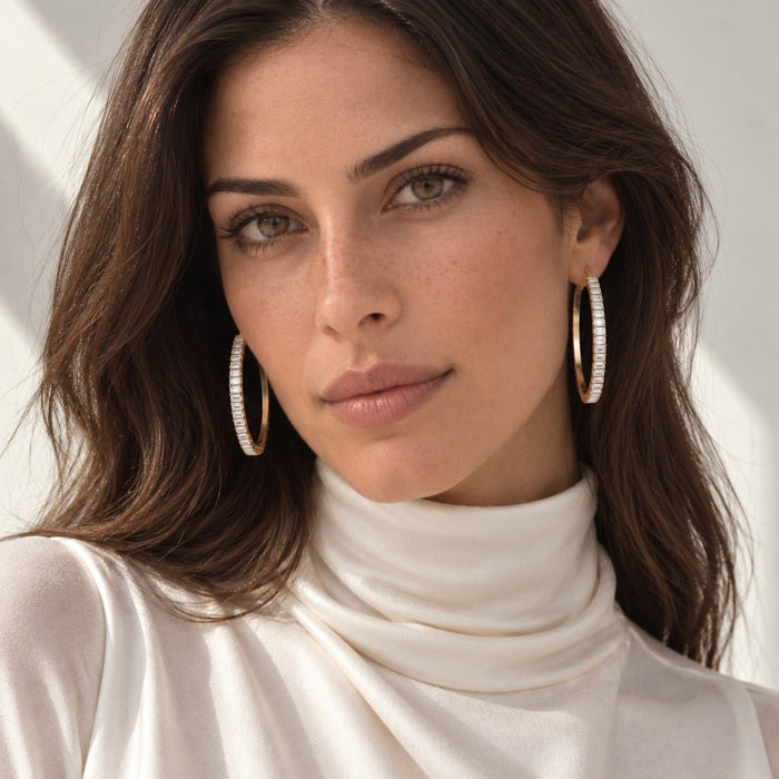 A close-up portrait of a woman wearing large silver-toned hoop earrings studded with small clear crystals, paired with a cream turtleneck top against a softly lit light background.