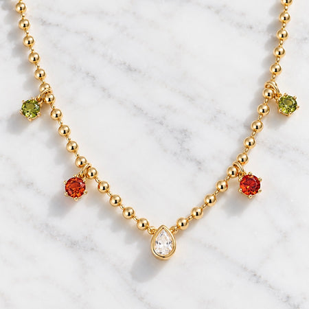 A gold beaded necklace with a teardrop-shaped clear gemstone pendant and three smaller round gemstone charms in green, orange, and red, displayed on a white marble background.