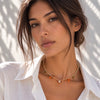 A close-up lifestyle shot of a woman wearing a delicate gold chain necklace with small green and orange charms and a central clear stone, styled with a white collared shirt against a sunlit white wall.