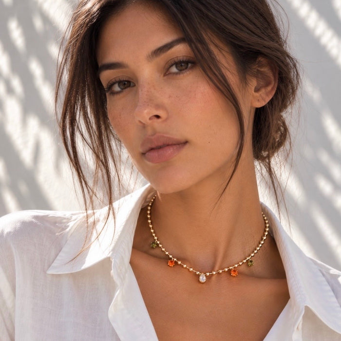 A close-up lifestyle shot of a woman wearing a delicate gold chain necklace with small green and orange charms and a central clear stone, styled with a white collared shirt against a sunlit white wall.