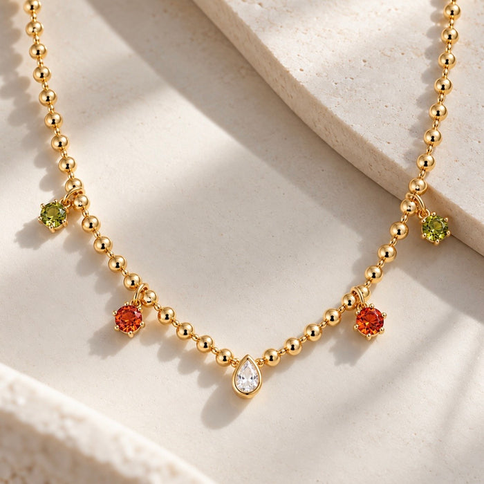 Gold beaded necklace with small dangling charms, including a central teardrop-shaped clear stone and three round colored stones in green, orange, and green, displayed on a light stone surface.