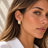 A close-up of a woman wearing a small, stacked wood bead earring. The earring features natural light wood tones, and the background shows soft, warm shadows on a light-colored wall.