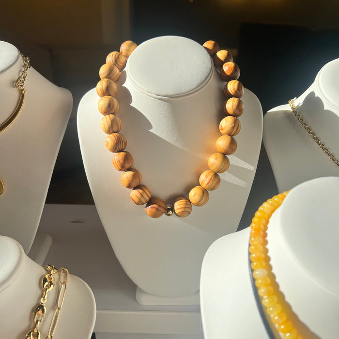Wooden beaded necklace on a white stand with gold chain necklace in the background.