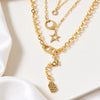 A gold layered necklace set displayed on a white surface with draped white fabric. The set includes a thick curb chain, a delicate chain with a crescent moon, a star, a safety pin, and a small shield charm, all embellished with small clear rhinestones.