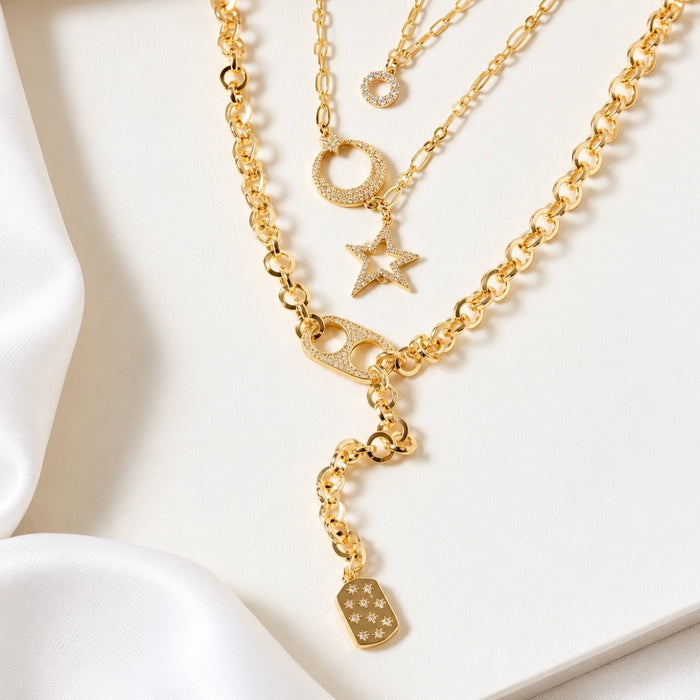 A gold layered necklace set displayed on a white surface with draped white fabric. The set includes a thick curb chain, a delicate chain with a crescent moon, a star, a safety pin, and a small shield charm, all embellished with small clear rhinestones.