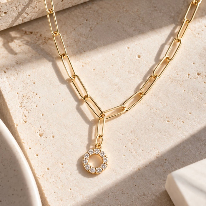 A gold paperclip chain necklace with a small circular pendant encrusted with clear stones, styled on a textured beige stone surface with soft shadows.