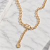A gold chain necklace with a Y-shaped design, featuring a small dangling shield charm embellished with clear crystals, displayed on a white marble surface.