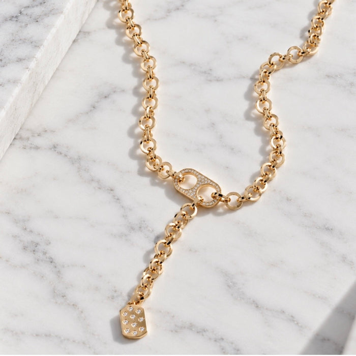 A gold chain necklace with a Y-shaped design, featuring a small dangling shield charm embellished with clear crystals, displayed on a white marble surface.