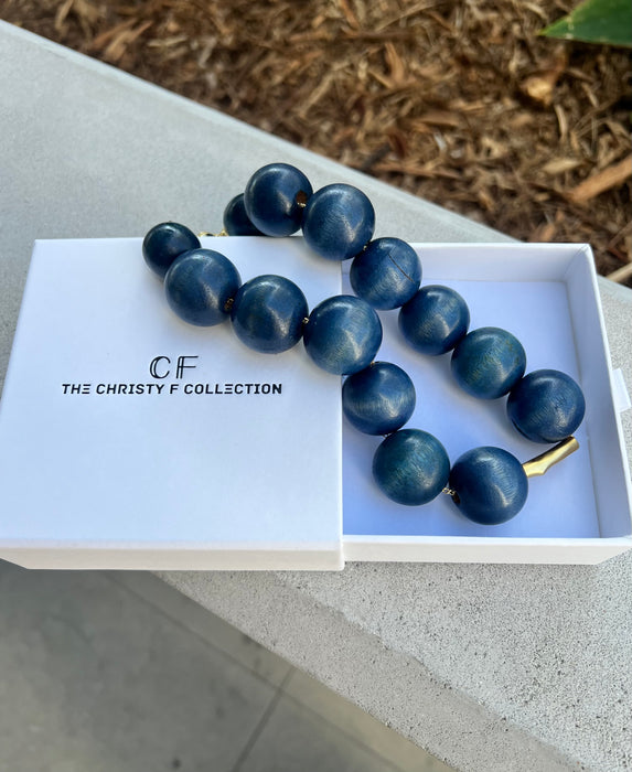 Blue beaded necklace in a white box with 'The Christy F Collection' branding on a concrete surface.