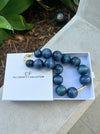 Blue beaded necklace in a white box with 'The Christy F. Collection' branding on a stone surface.
