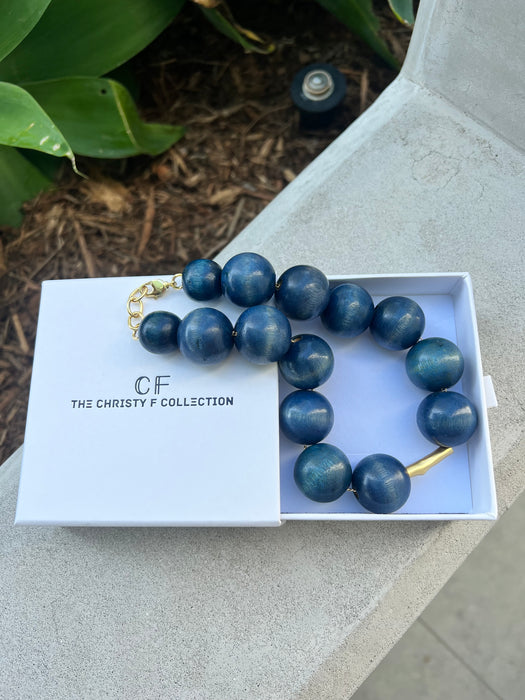 Blue beaded necklace in a white box with 'The Christy F. Collection' branding on a stone surface.