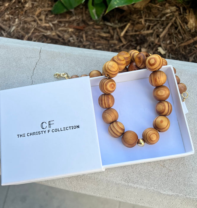 Wooden necklace in an open box with 'The Christy F Collection' branding on a concrete surface.