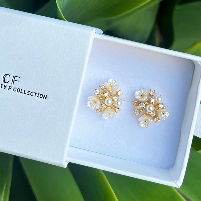 A pair of gold-filled crystal flower stud earrings displayed in an open white box with the brand 'OF' visible on the box's lid, set against a leafy background.