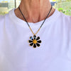 Person wearing a necklace with a black and gold flower pendant on a white shirt.