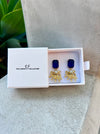 Gold earrings with blue stones in a white box labeled 'The Christy F Collection' on a stone surface.