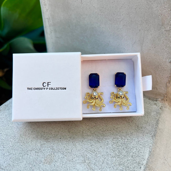 Gold earrings with blue stones in a white box labeled 'The Christy F Collection' on a stone surface.