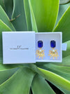 Gold and blue earrings in a white box with 'CF' branding on a green leafy background