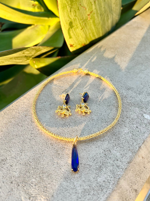 Gold necklace with blue pendant and matching earrings on a stone surface with green leaves in the background