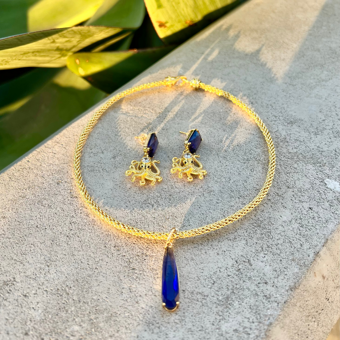Gold necklace with blue pendant and matching earrings on a stone surface with green leaves in the background