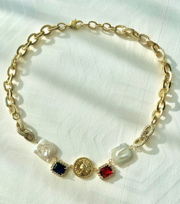 A gold-colored chunky chain necklace featuring white freshwater pearls and red ruby gemstones, displayed on a white fabric surface.