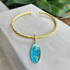 Gold bracelet with a blue stone on a textured surface with green plants in the background