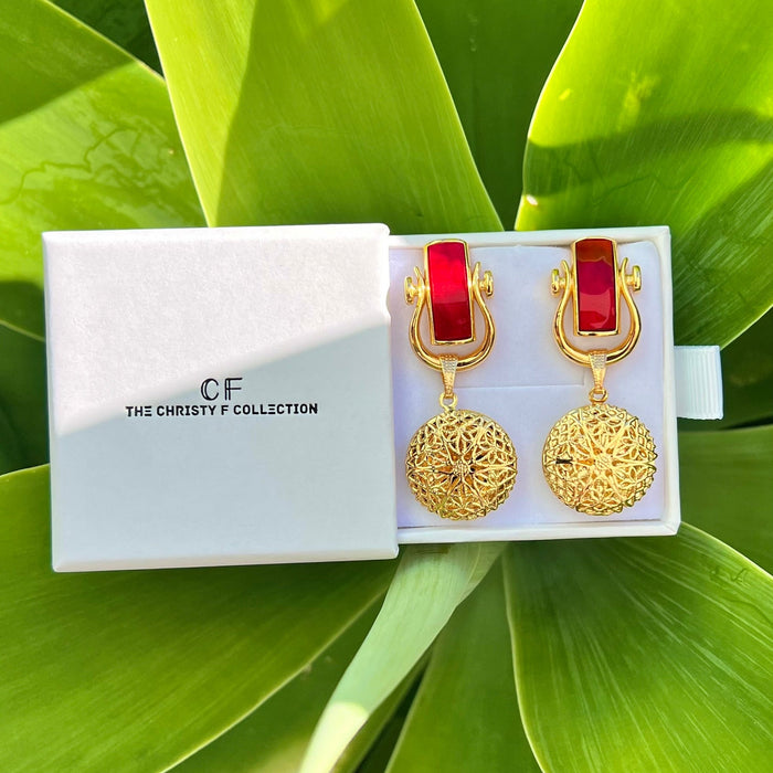 Gold earrings with red accents in a white box labeled 'The Christy F Collection' on green leaves.