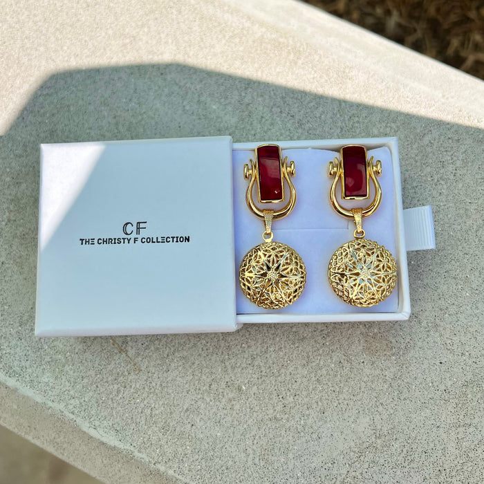 Gold earrings with red stones in a 'The Christy F Collection' box on a textured surface.
