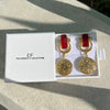 Gold and red earrings in a white box on a textured surface with natural light.