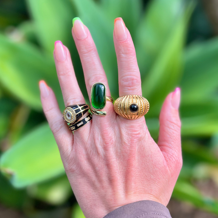 Hand wearing three rings with green and black stones against a blurred green leafy background