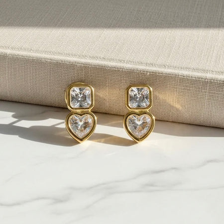 Gold heart-shaped earrings with clear gemstones on a textured surface