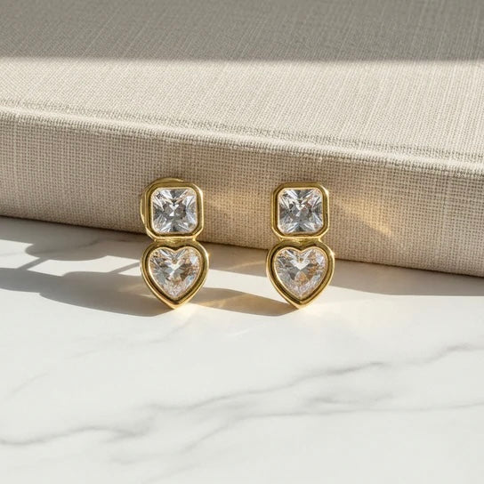 Gold heart-shaped earrings with clear gemstones on a textured surface