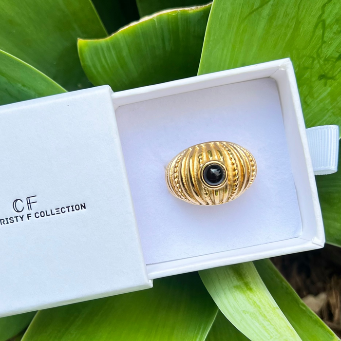 Gold ring with black gemstone in a white box labeled 'CF Collection' on green leaves.