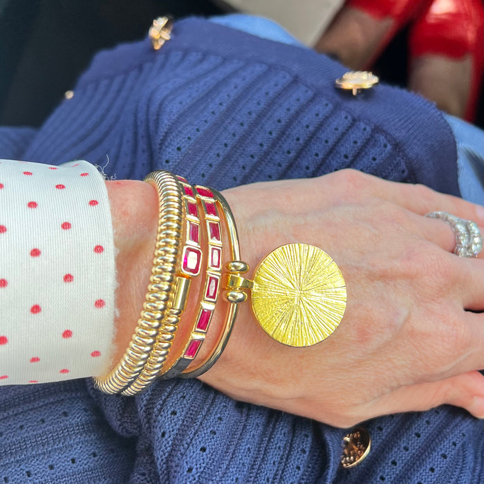 A close-up of a person's wrist wearing multiple stacked bracelets, including a large textured gold disc bracelet, a gold coiled bracelet, and red-and-gold striped bangles, paired with a white polka-dot shirt cuff and a blue knit sweater.