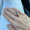 Hand wearing a gold ring with blue and red gemstones on a white background
