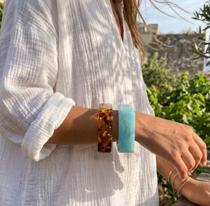 Person wearing two bracelets, one brown and one blue, with a blurred outdoor background.