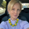 Woman wearing a purple polka dot blouse and yellow beaded necklace sitting in a car.